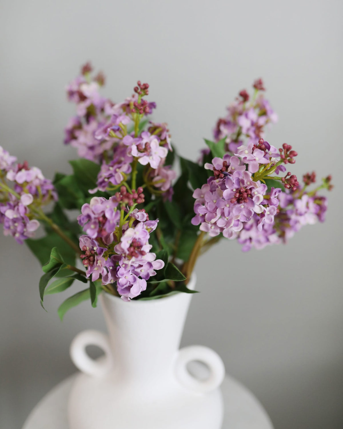 Bouquet of purple artificial lilac in a white vase on a gray background