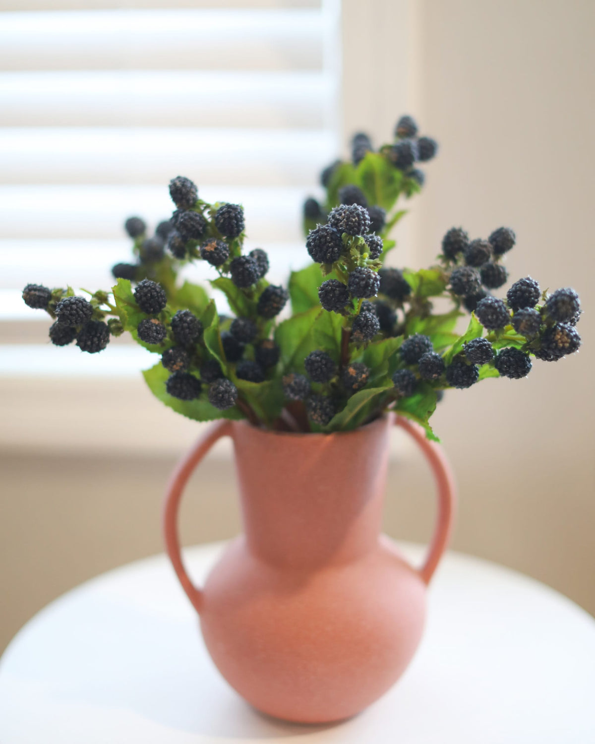 A terracotta-colored ceramic pitcher vase with two handles, filled with artificial blackberries and green leaves, set on a white round table in front of a softly blurred window with white blinds.