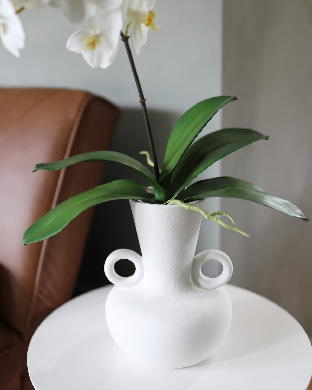 A white ceramic vase with two loop handles sits on a round white side table, holding a green leafy plant with white orchid flowers. A brown leather chair is visible in the background against a neutral gray wall.
