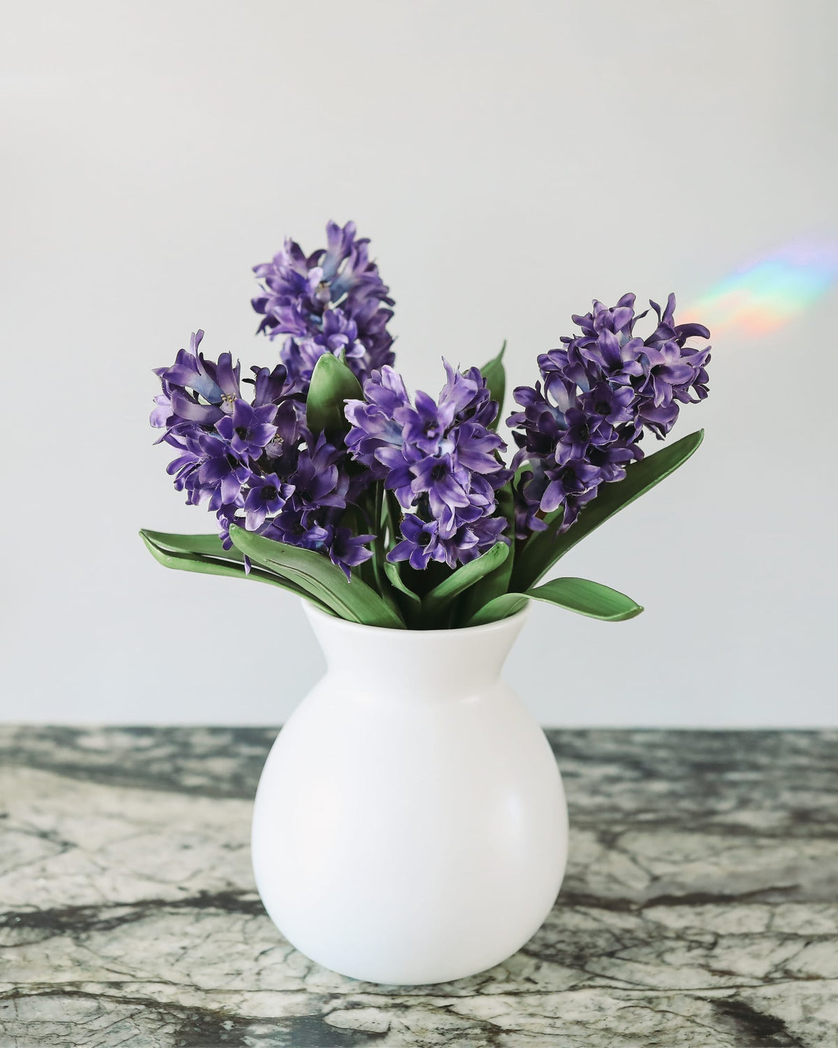 Purple hyacinth artificial flowers arranged in a white ceramic vase, set on a gray marble-patterned surface against a light gray background.
