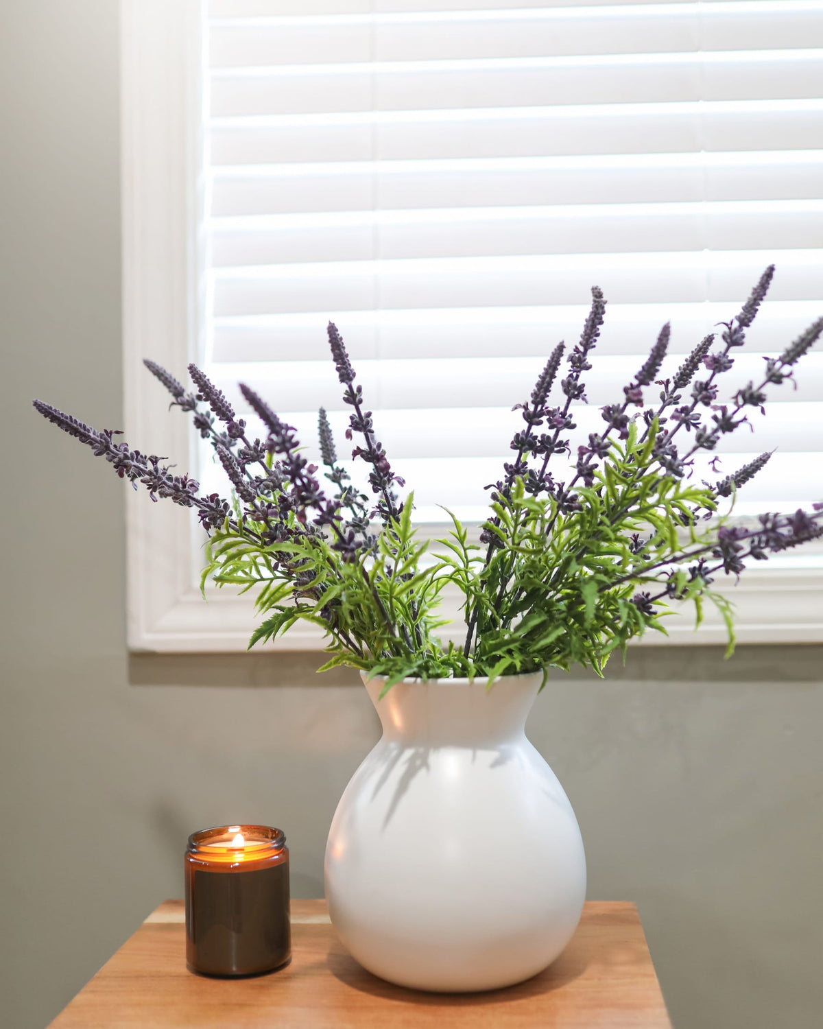 A white ceramic vase filled with faux purple lavender stems, placed on a wooden table next to a lit candle in front of a window with white blinds.