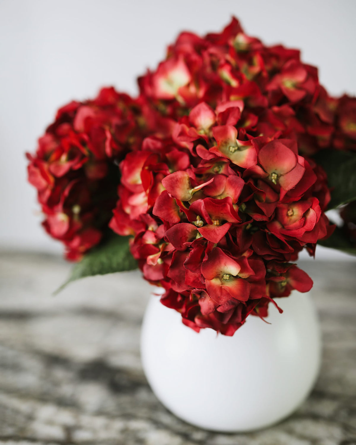 A bouquet of deep red faux hydrangea flowers with green leaves arranged in a round white ceramic vase, set on a rustic gray wooden surface against a soft light gray background.