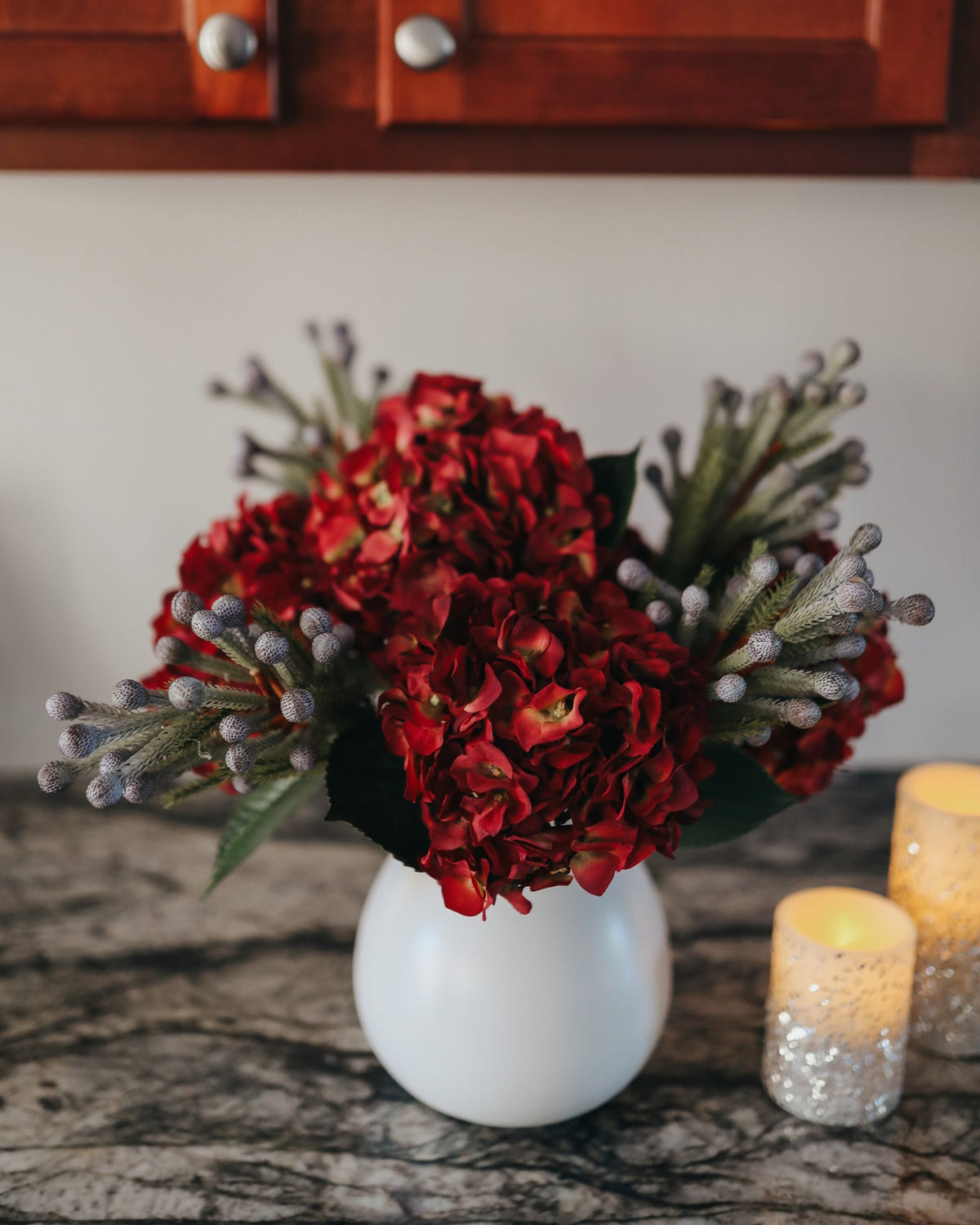 An artificial floral arrangement of deep red hydrangea-like blooms with gray-green textured stems in a round white vase, set on a dark marble surface beside two lit candles.