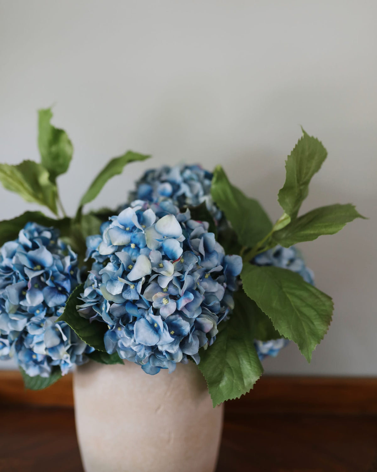 Blue hydrangea artificial flowers with green leaves arranged in a light beige ceramic vase on a wooden surface against a neutral gray wall.