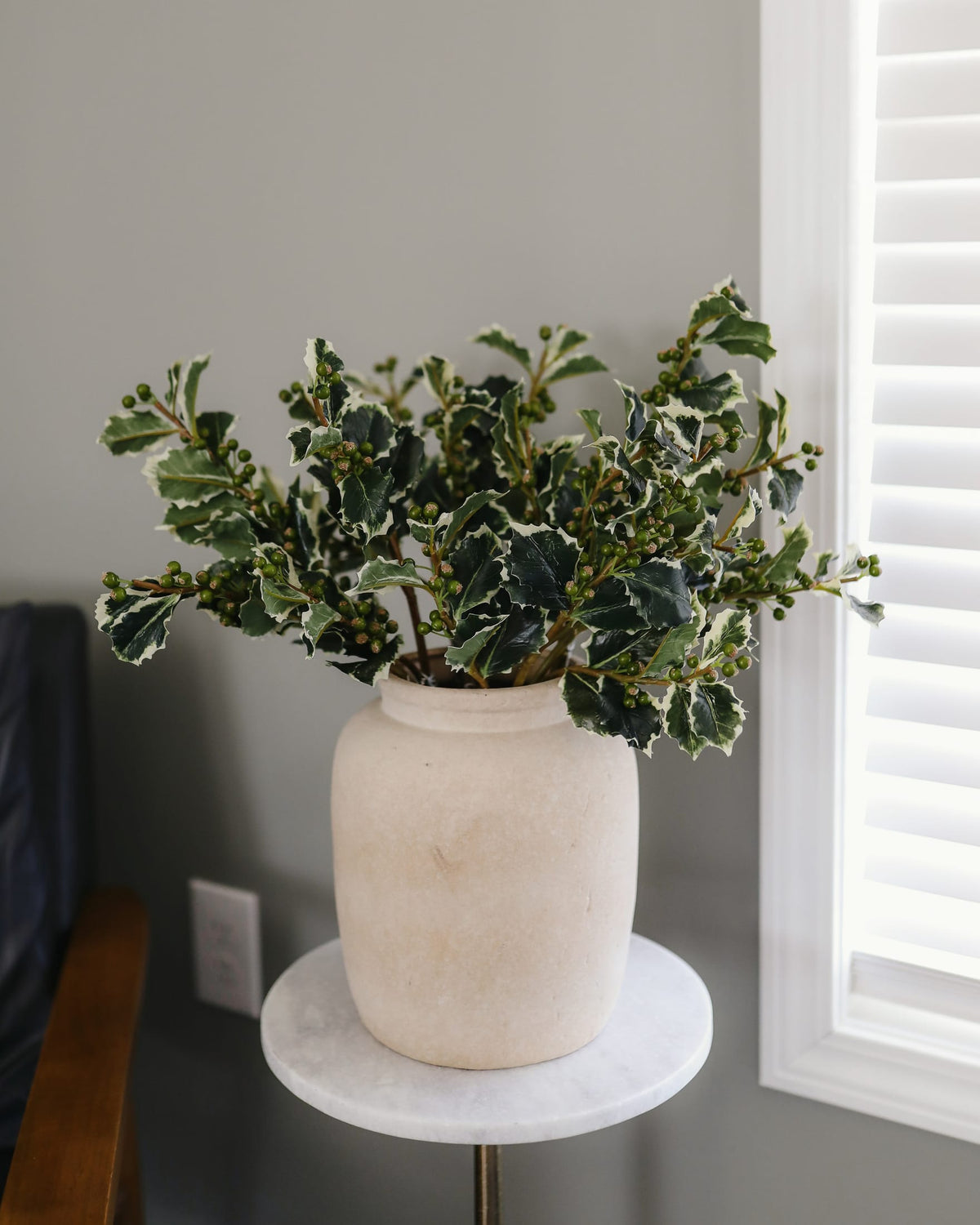 A potted variegated holly plant with green and white leaves, arranged in a textured cream-colored ceramic pot on a small round white marble side table. The table is set against a gray wall next to a window with white shutters.