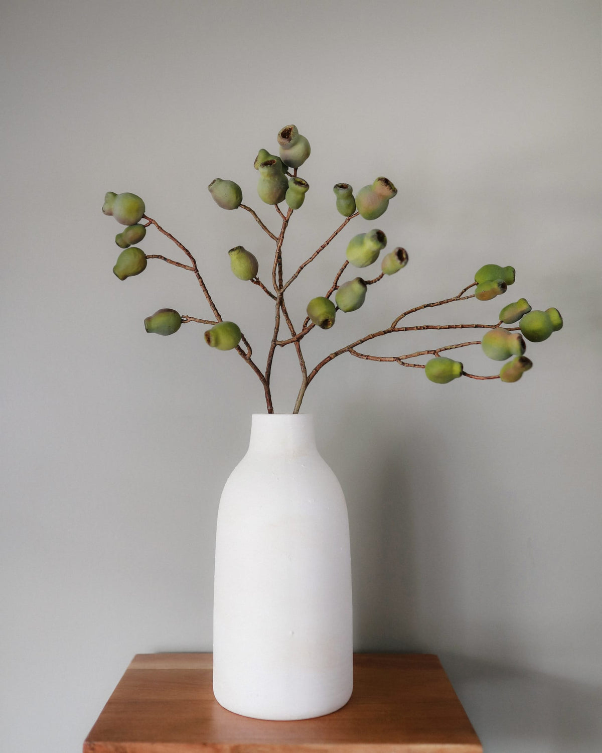 A white ceramic bottle-shaped vase holding artificial Eucalyptus Pods with small green buds, displayed on a wooden surface against a plain light gray wall.