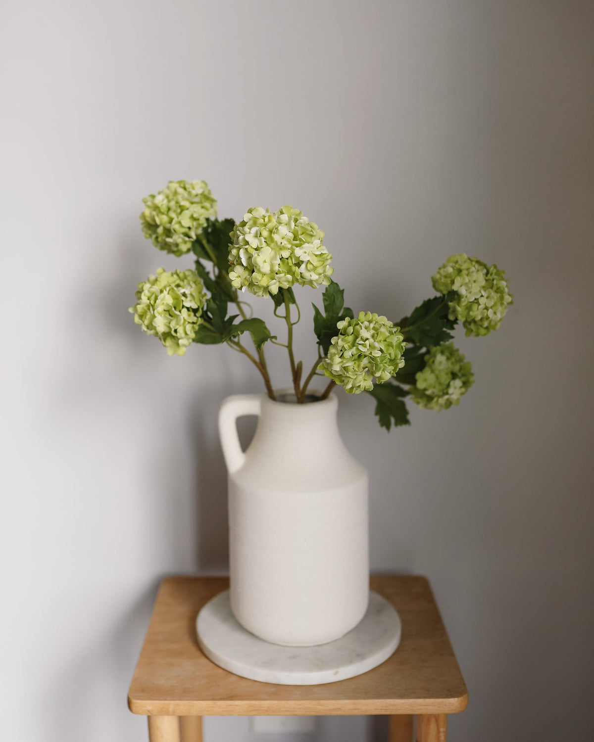 White vase with artificial Green Snowball flowers on a wooden table against a gray wall