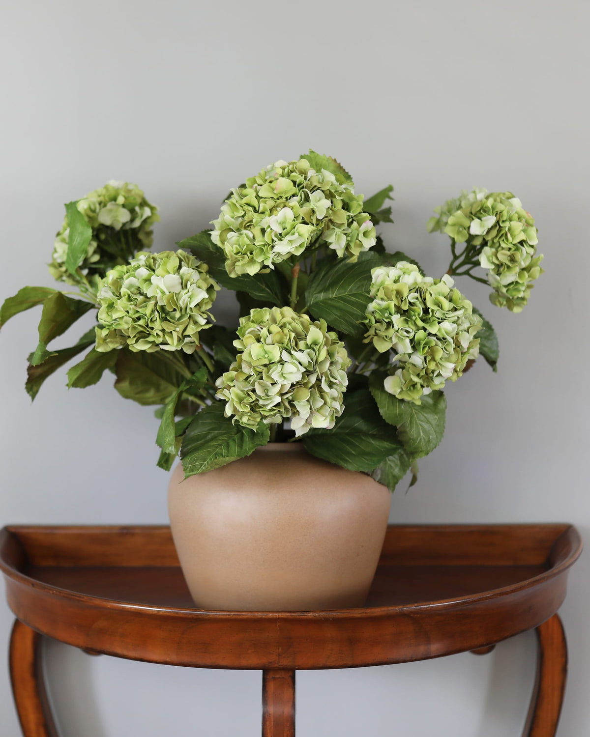 A faux green hydrangea floral arrangement in a matte beige ceramic pot, displayed on a curved wooden side table against a plain light gray wall.