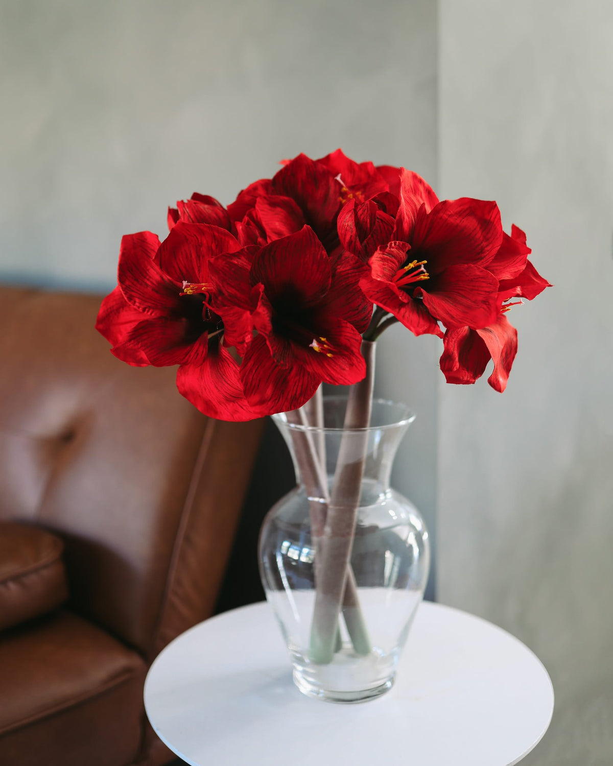 A bouquet of vibrant red faux amaryllis in a clear glass vase, displayed on a small white side table next to a brown leather armchair against a textured gray wall.