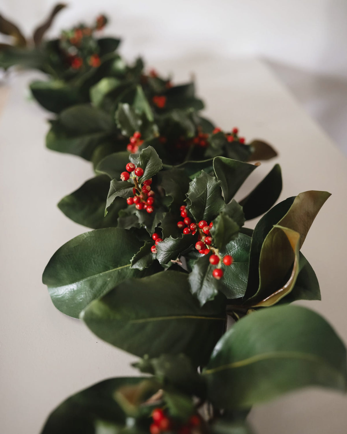 A green garland with glossy leaves and clusters of small red holly berries arranged diagonally on a light neutral surface.