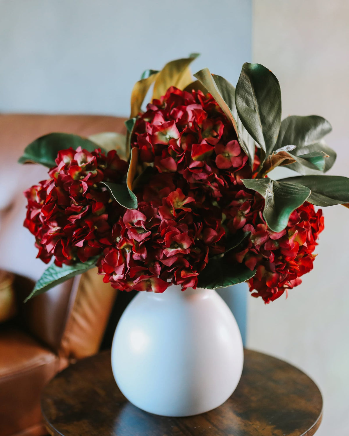 A bouquet of deep red faux hydrangea flowers with green leaves arranged in a matte white ceramic vase, set on a dark wooden table in a softly lit indoor setting.