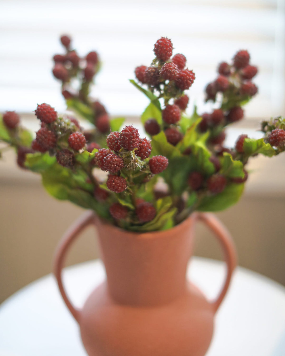 Artificial raspberries arranged in a terracotta-colored ceramic vase with two handles, set on a white surface.
