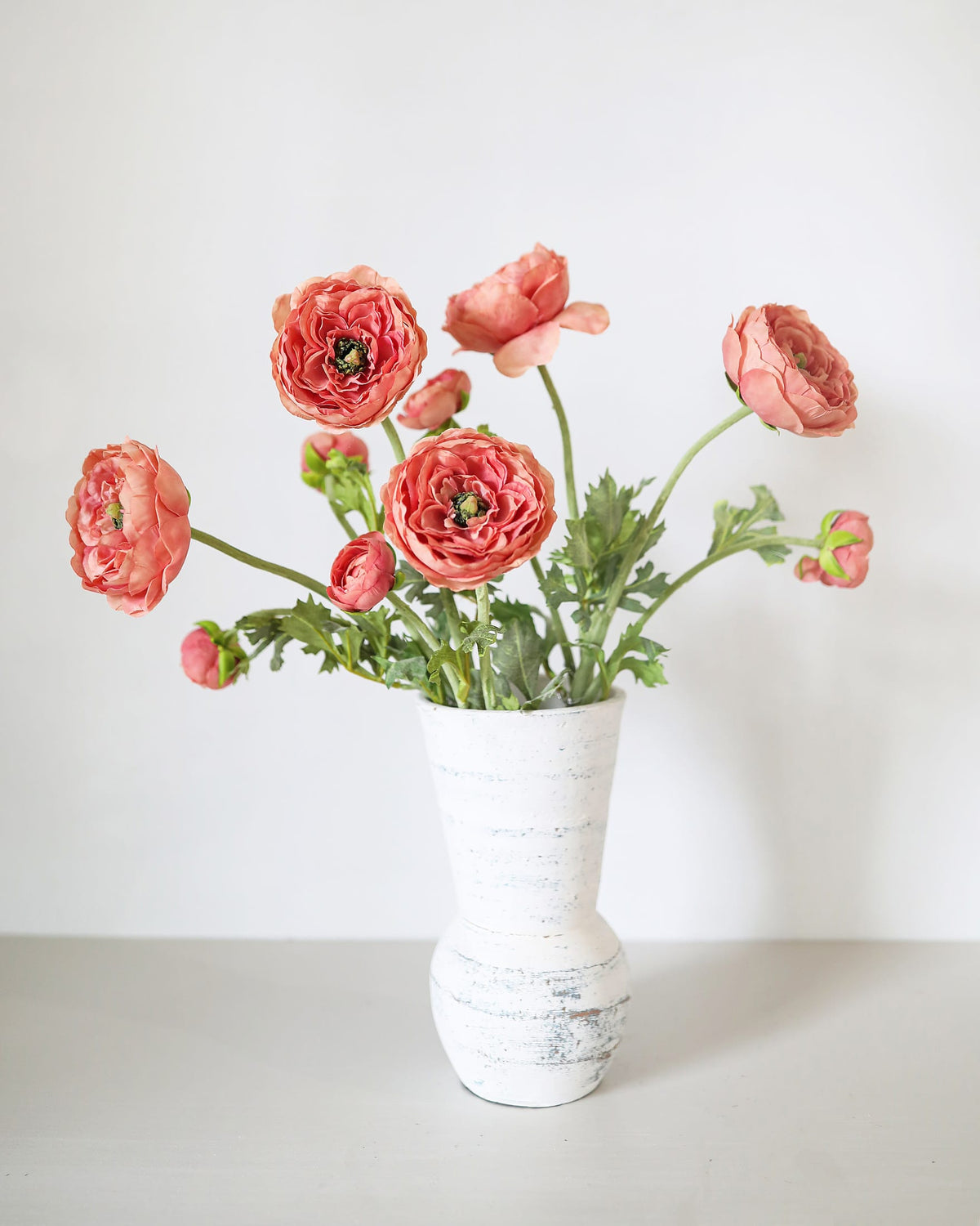 A bouquet of coral-pink faux ranunculus flowers with green stems and leaves, arranged in a distressed white ceramic vase against a clean white background.