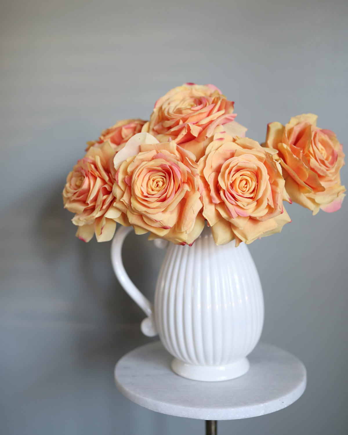A bouquet of peach and orange faux roses arranged in a white ribbed ceramic pitcher vase, set on a round white marble side table against a soft gray background.