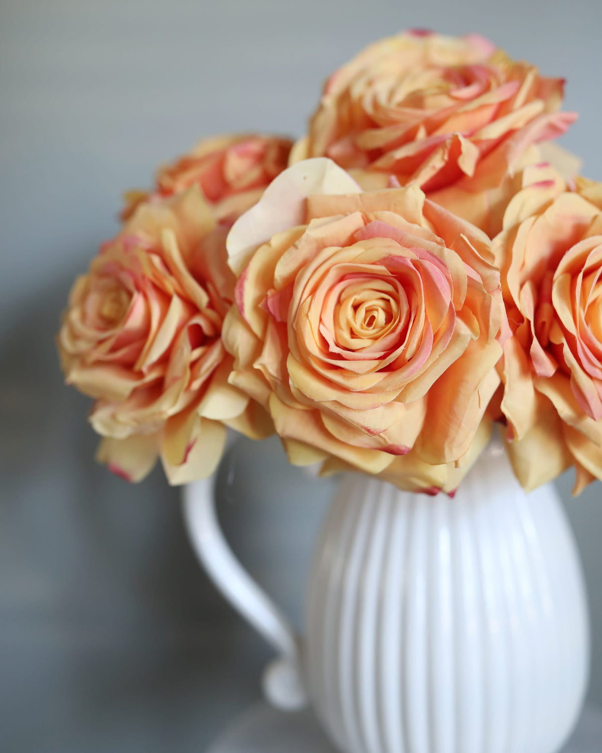 A bouquet of peach and pink faux roses arranged in a white ribbed ceramic pitcher against a soft gray background.
