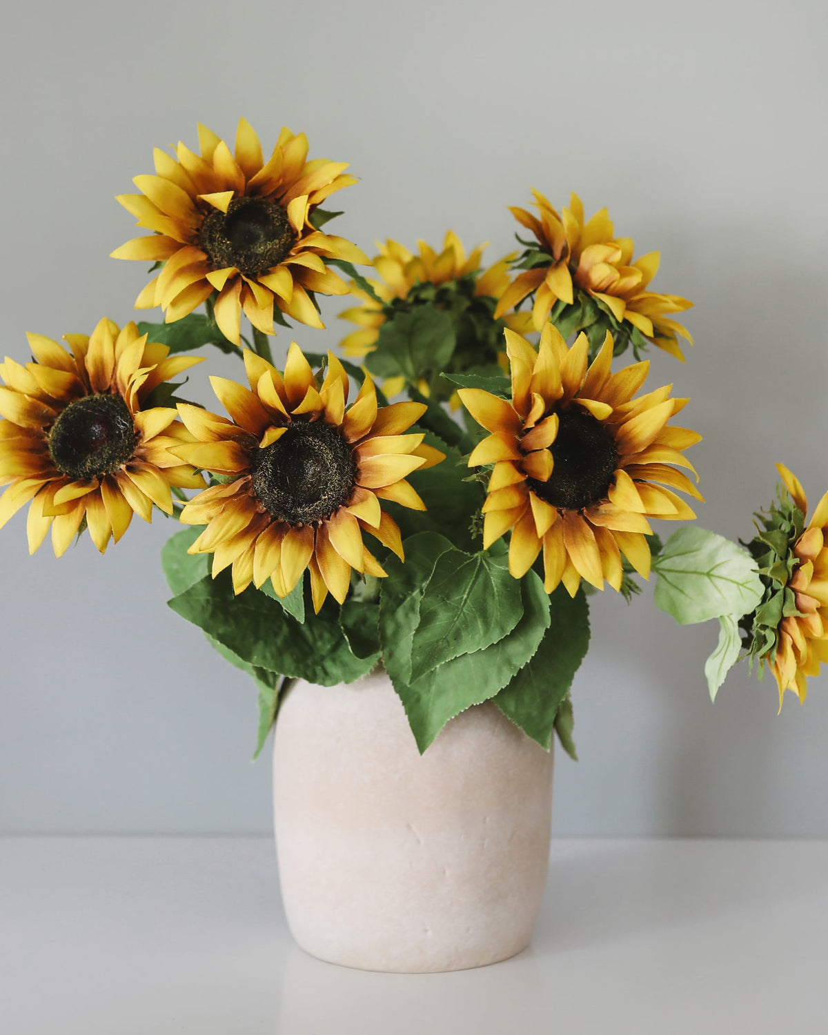 A bouquet of artificial yellow sunflowers with dark brown centers and green leaves, arranged in a matte off-white ceramic vase against a plain light gray background.