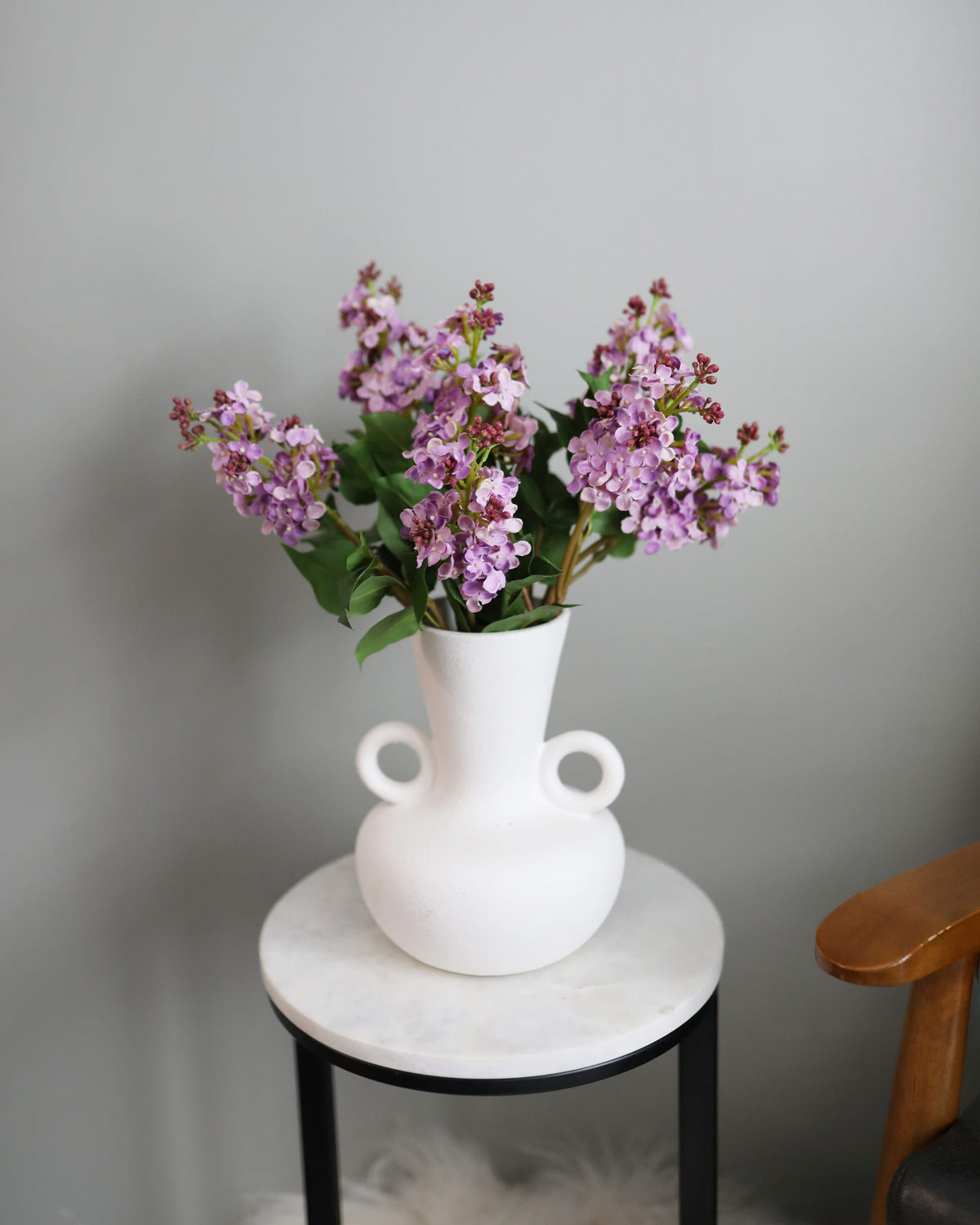 A white ceramic vase with two side loops, filled with faux light purple lilac flowers and green leaves, displayed on a small marble-top side table against a plain gray wall.