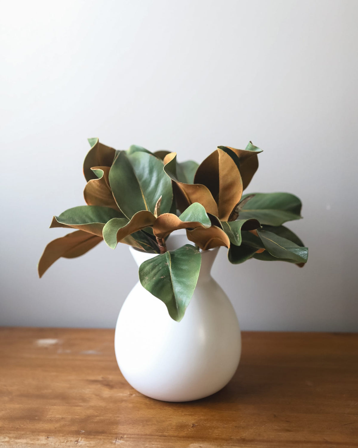 A white ceramic vase holding a faux magnolia  arrangement with glossy green and brown leaves, set on a wooden surface against a plain light gray wall.