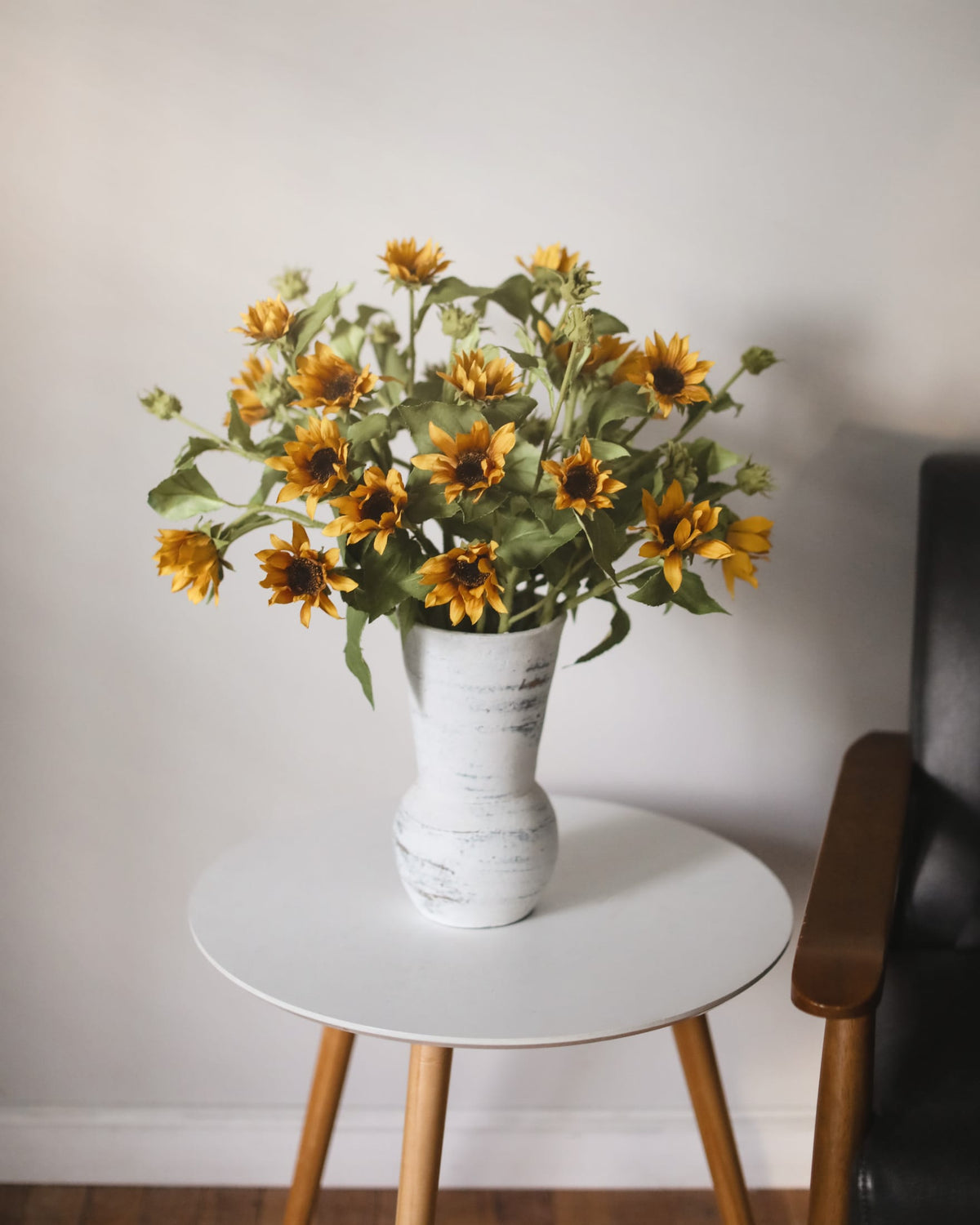 White vase with faux sunflowers on a round white table against a plain wall.