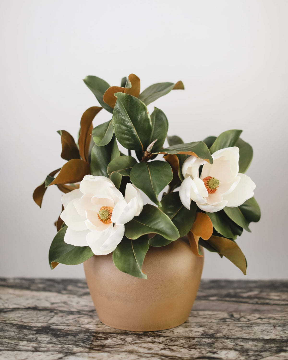 A potted artificial magnolia plant with white blossoms, green leaves, and brown foliage, arranged in a round beige ceramic pot on a gray marble surface against a light gray background.