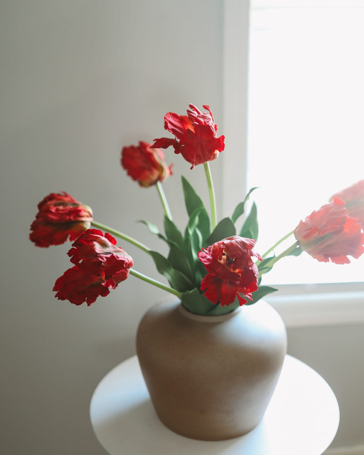 Red faux tulips with green leaves arranged in a matte beige ceramic vase, placed on a white round table near a bright window.