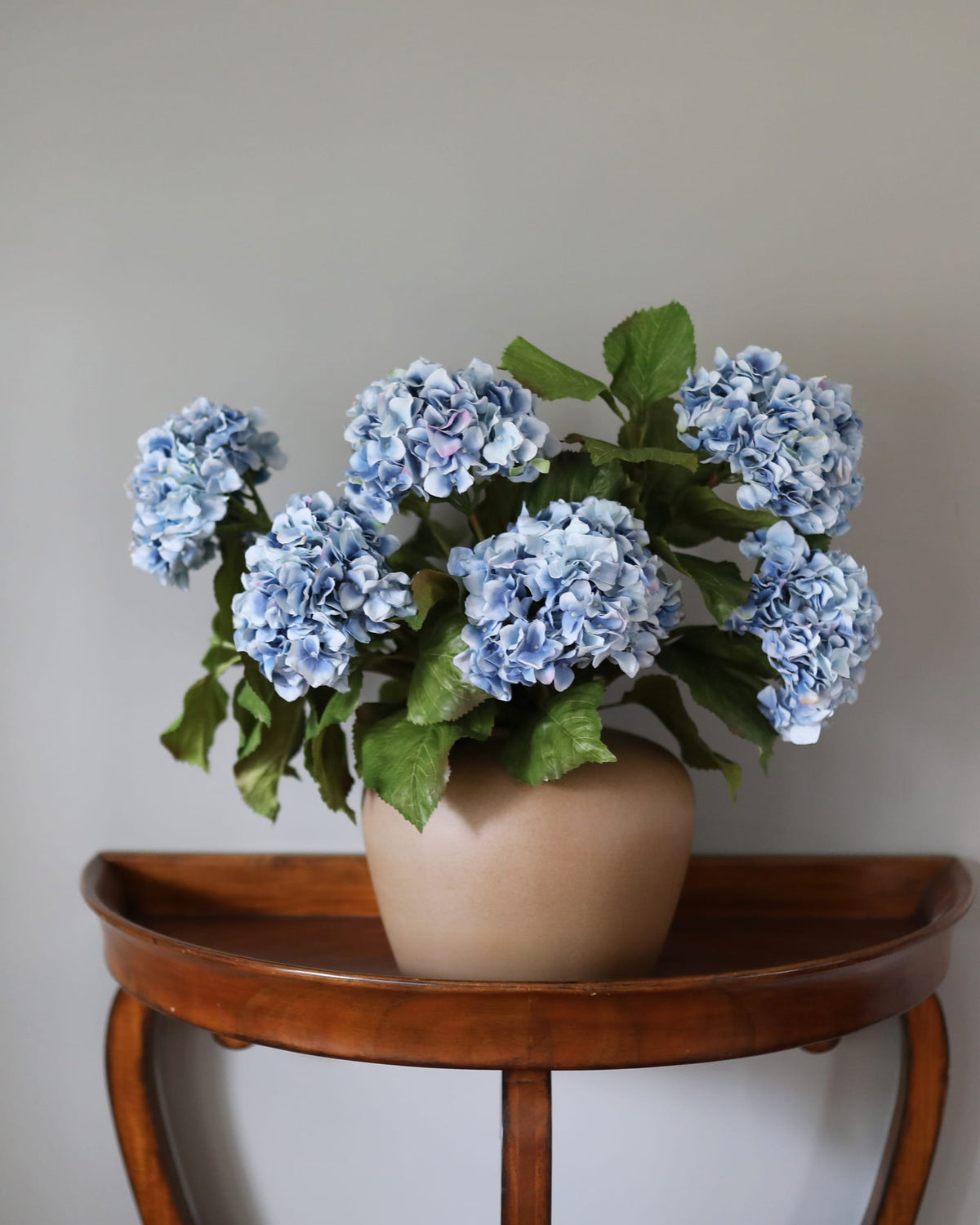 A potted faux artificial hydrangea with light blue flowers and green leaves, displayed in a beige ceramic pot on a curved wooden side table against a plain light gray wall.