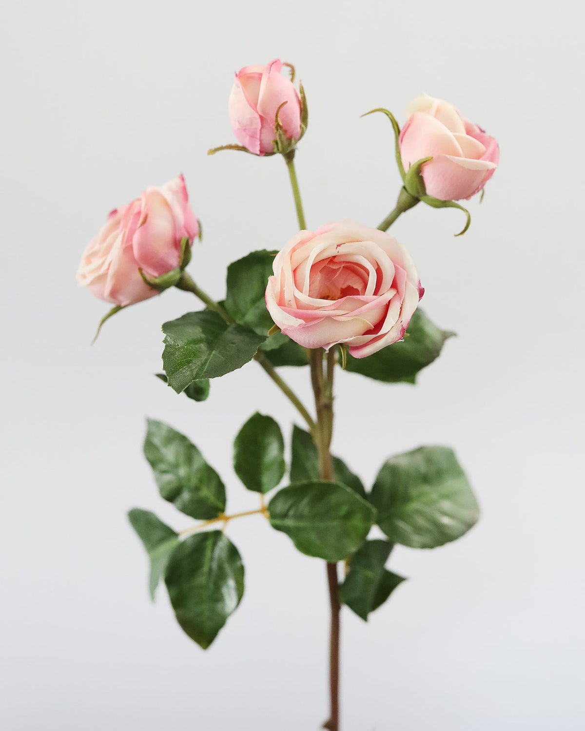 A single artificial rose stem with four blooms, featuring pale pink and white petals, green leaves, and brown stems, set against a plain light gray background.
