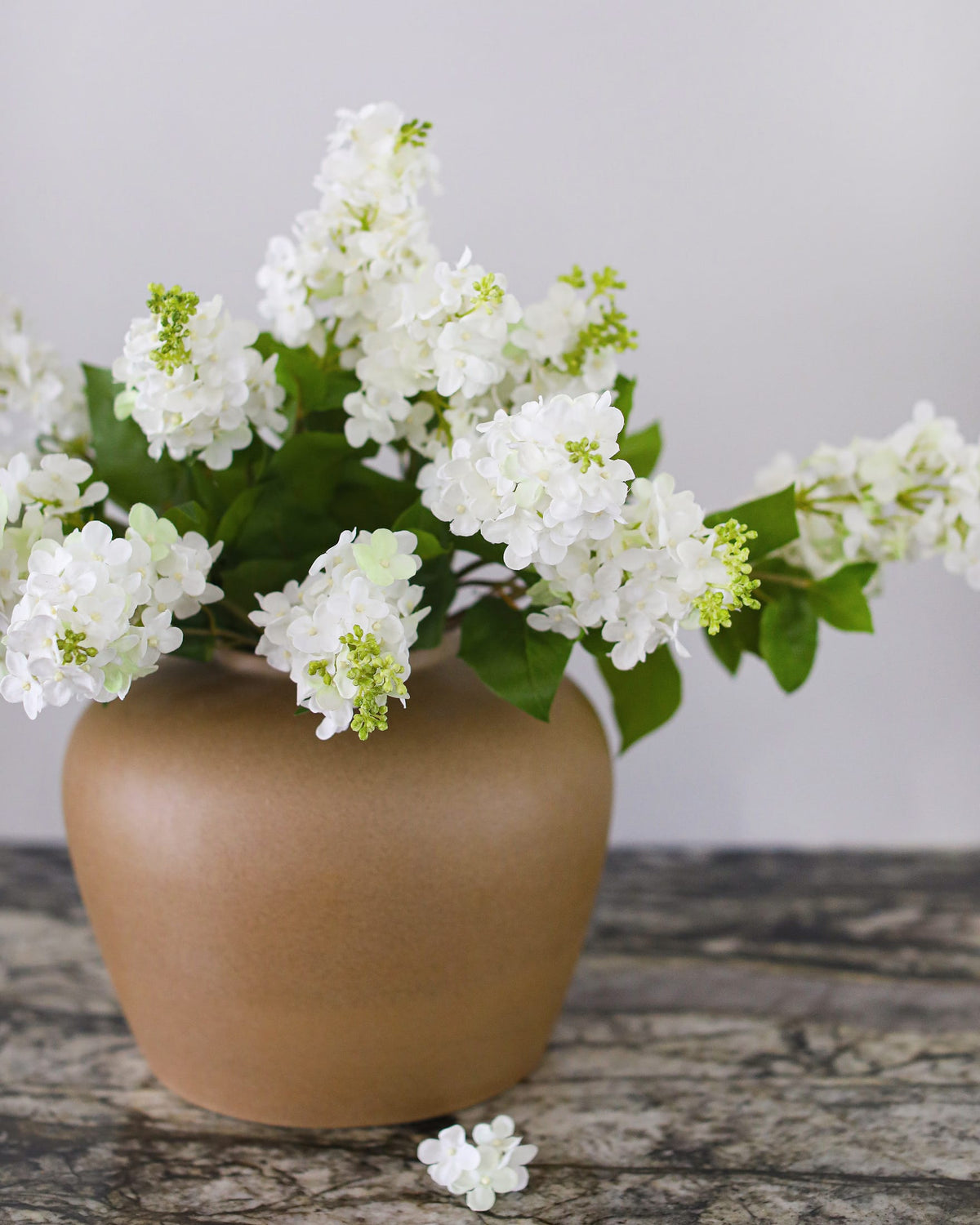 White lilac artificial flowers with green leaves arranged in a matte brown ceramic vase on a rustic wooden surface against a light gray background.