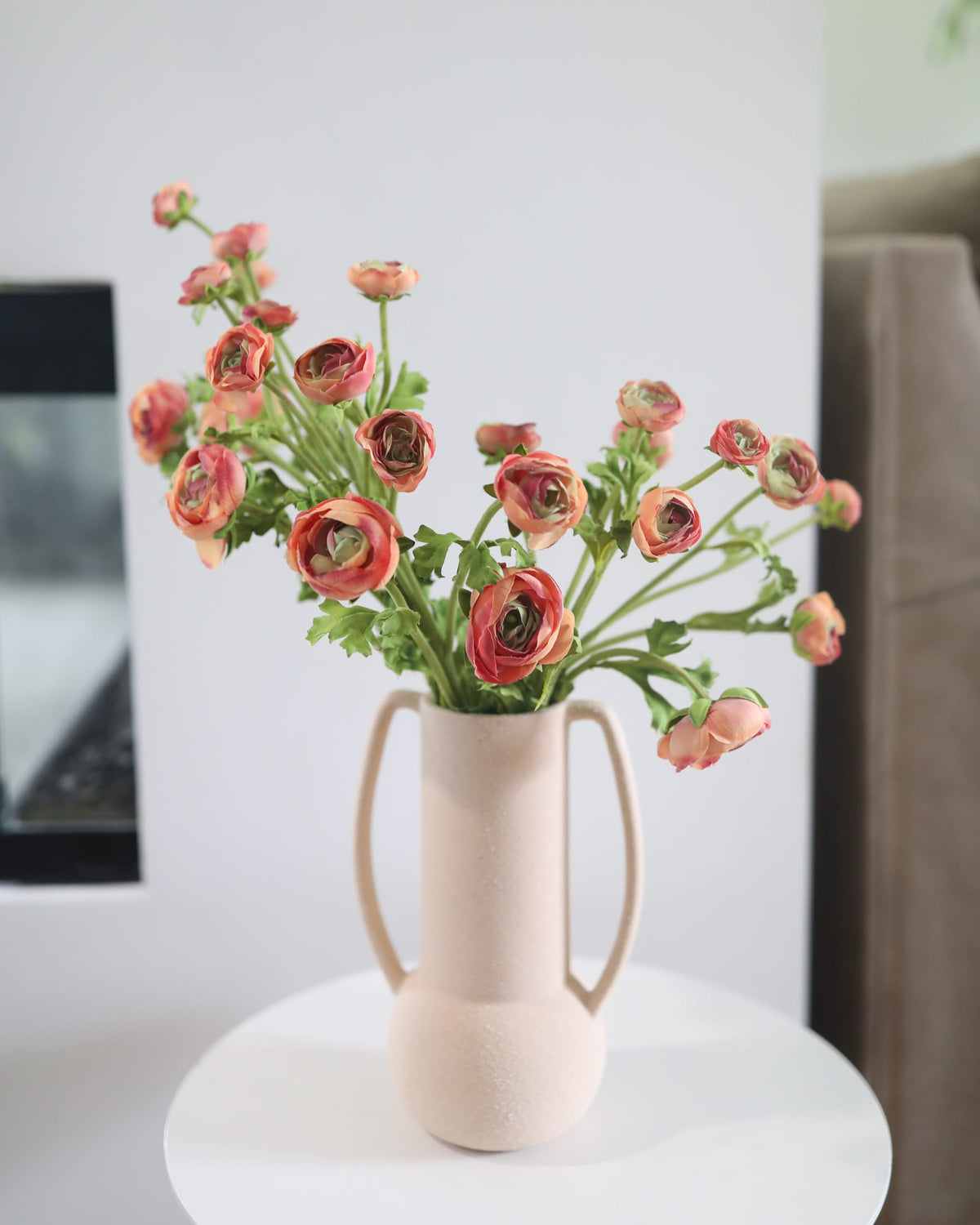 Beige ceramic vase with faux pink ranunculus on a white surface