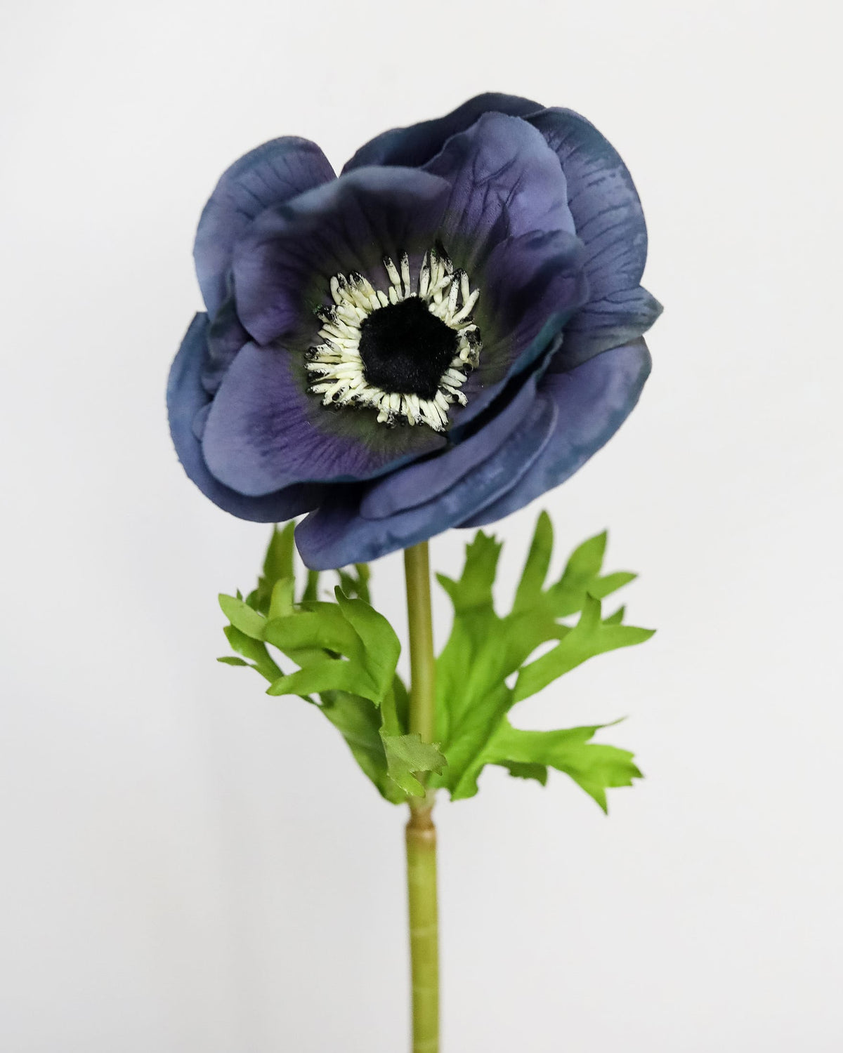 A single artificial anemone flower with dark blue-purple petals, a black and white center, and green leaves on a stem, photographed against a plain light gray background.
