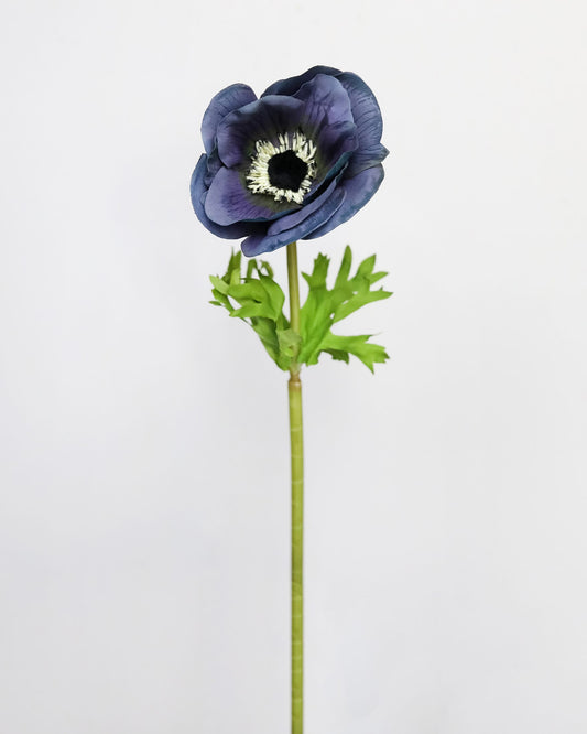 A single artificial anemone flower with dark blue-purple petals, a black and white center, and green ruffled leaves on a long stem, photographed against a plain light gray background.