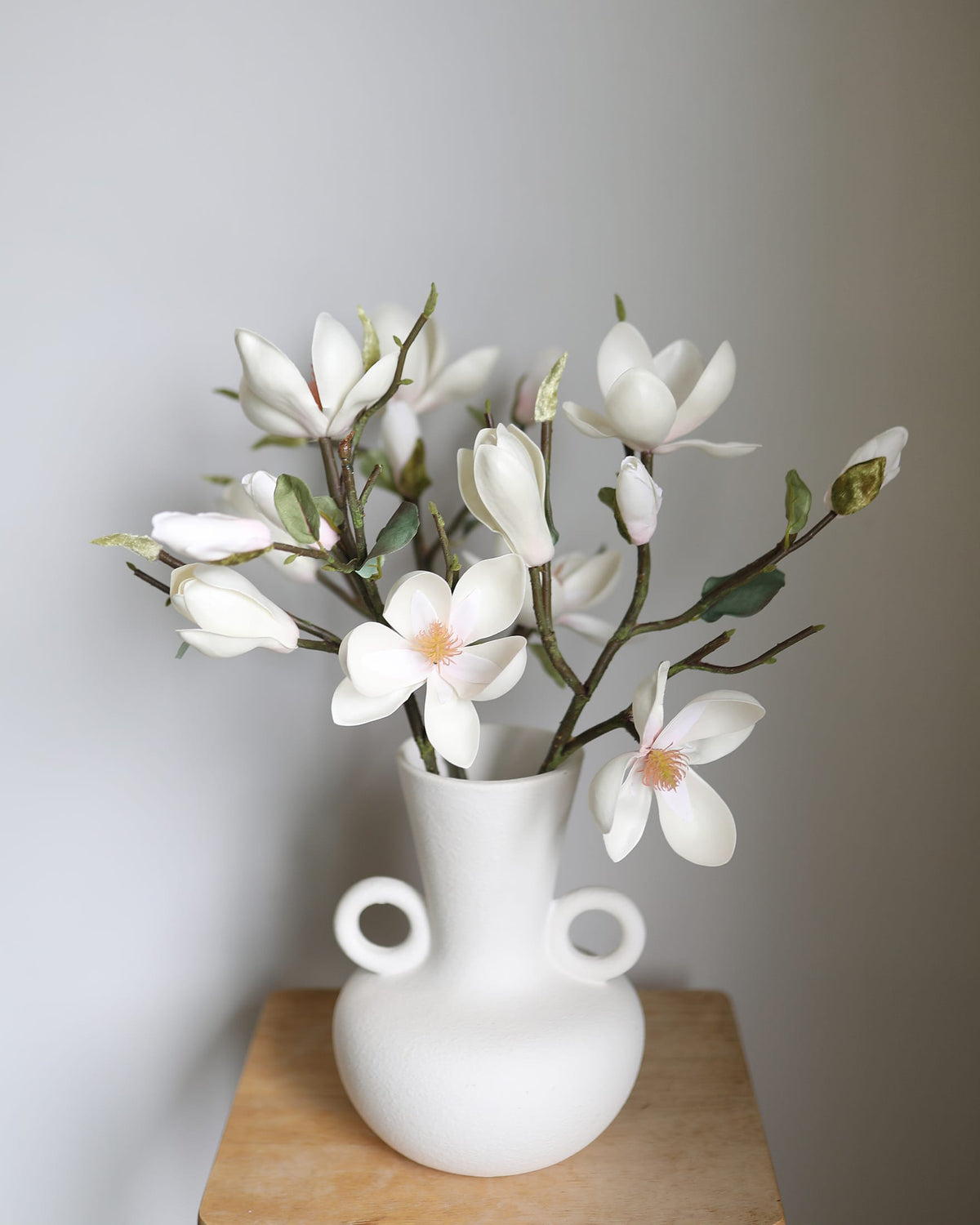 White vase with white faux magnolia on a wooden surface against a gray wall