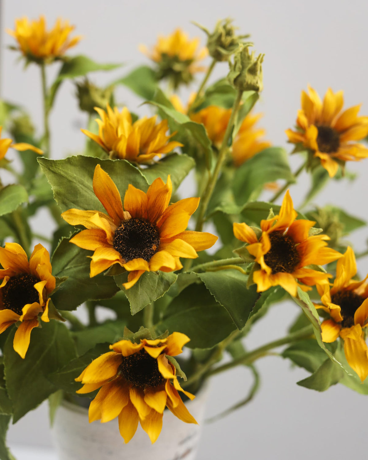 A bouquet of artificial yellow sunflowers with dark brown centers and green leaves, arranged in a white pot against a light gray background.