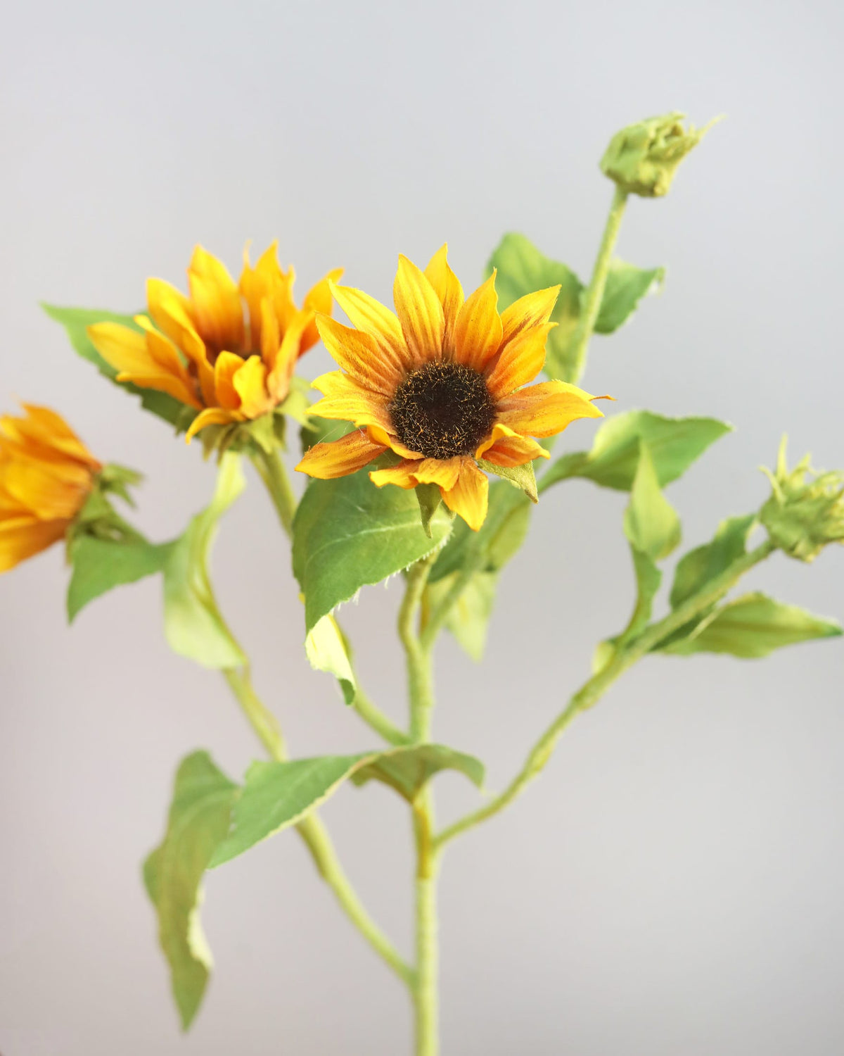 Artificial sunflower stem with three yellow-orange blooms, green leaves, and buds on a light gray background.