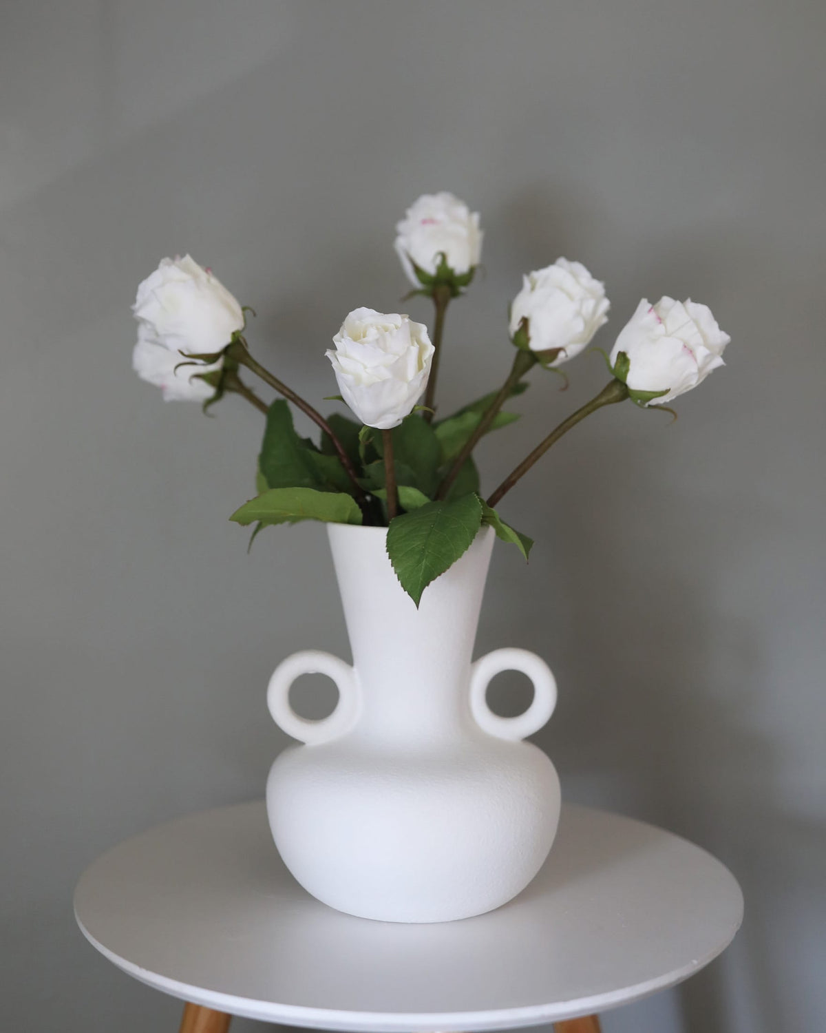 A matte white ceramic vase with two side loops, holding white faux roses with green leaves, set on a round white table against a neutral gray background.
