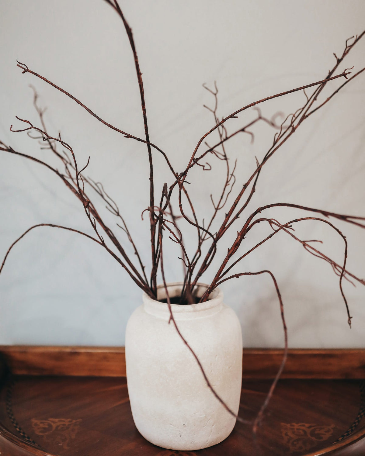 White vase with fake branches on a wooden surface against a light wall.