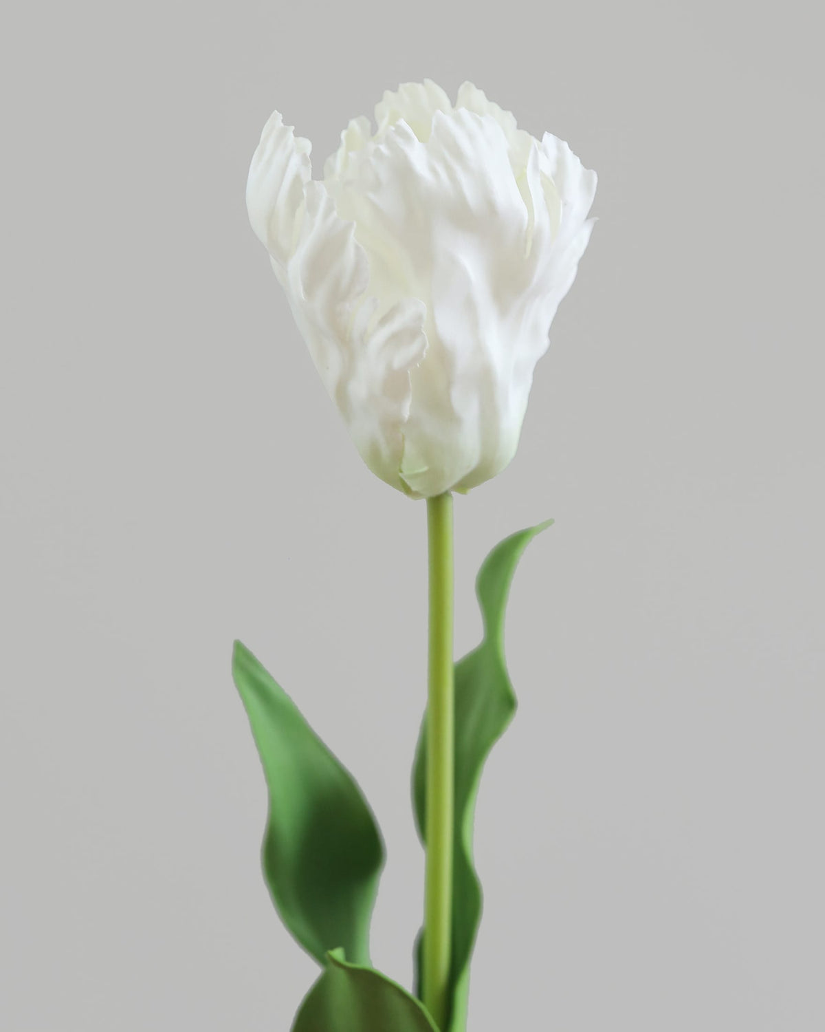 A single white faux tulip flower with a long green stem and leaves, centered against a plain light gray background.