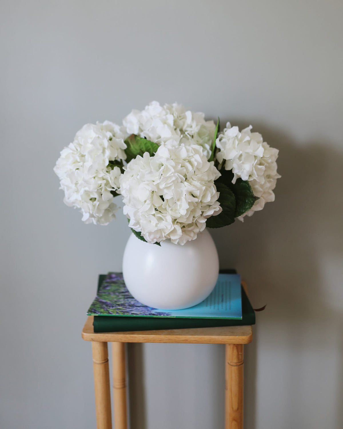 White faux real touch hydrangeas in a vase on a small wooden table against a plain wall.