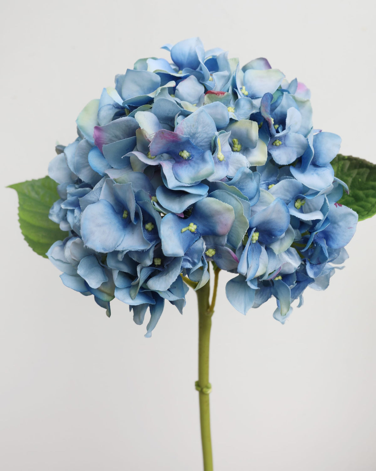 A single artificial hydrangea stem with a large round cluster of light blue to periwinkle-blue flowers, accented with green leaves and a green stem, set against a plain light gray background.