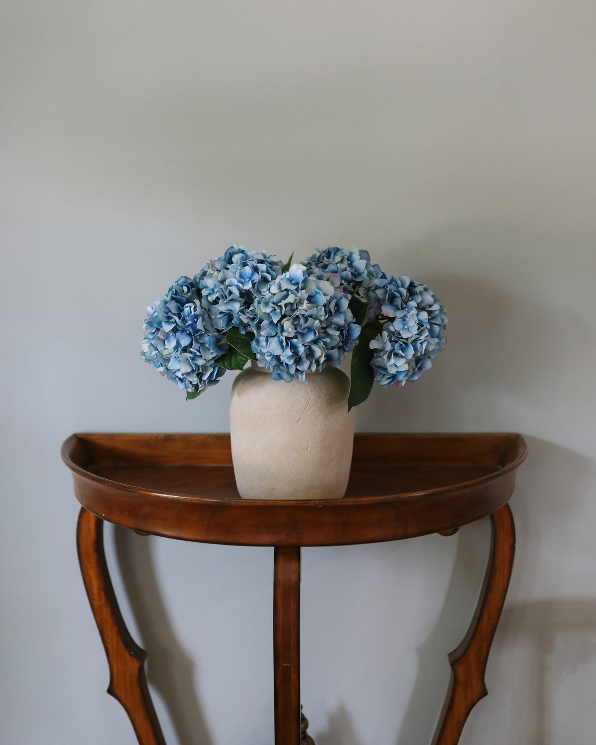 Artificial Blue hydrangea flowers arranged in a beige ceramic vase, placed on a curved wooden console table against a plain light gray wall.
