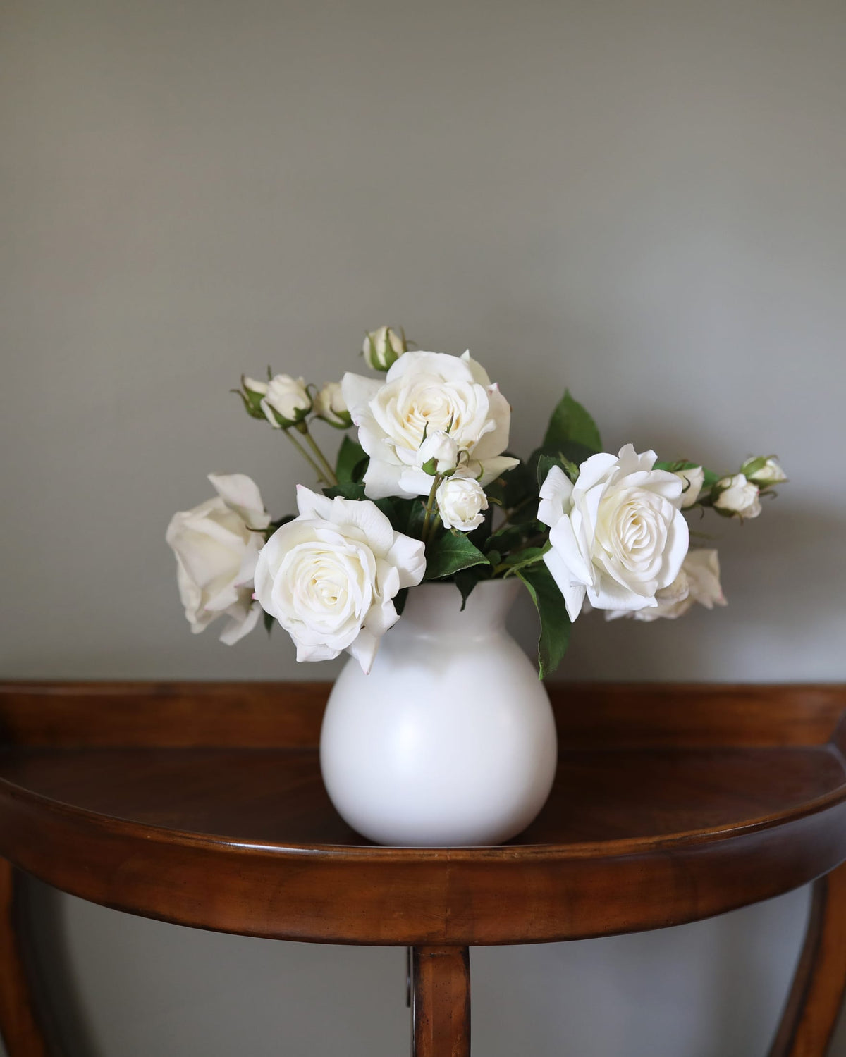 White faux roses arranged in a matte white ceramic vase, set on a curved wooden console table against a neutral gray wall.