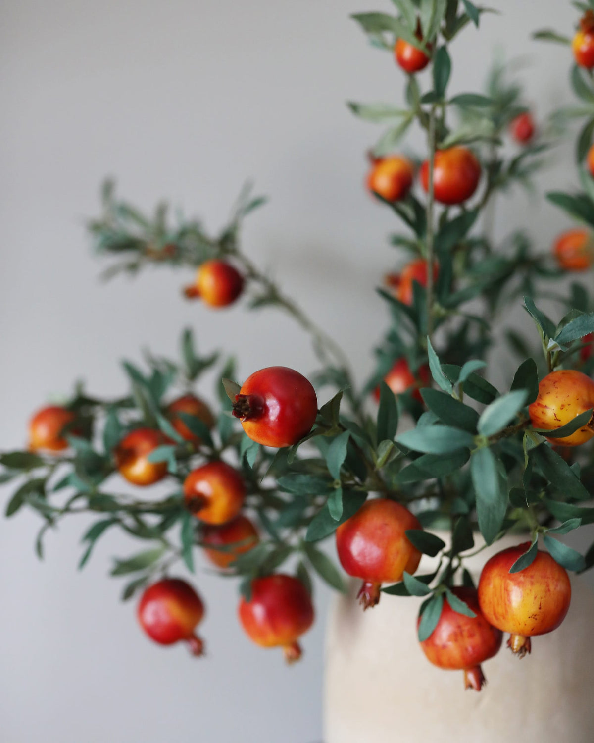 Artificial pomegranate branches with glossy red-orange fruits and green leaves arranged in a light beige ceramic vase against a soft neutral background.