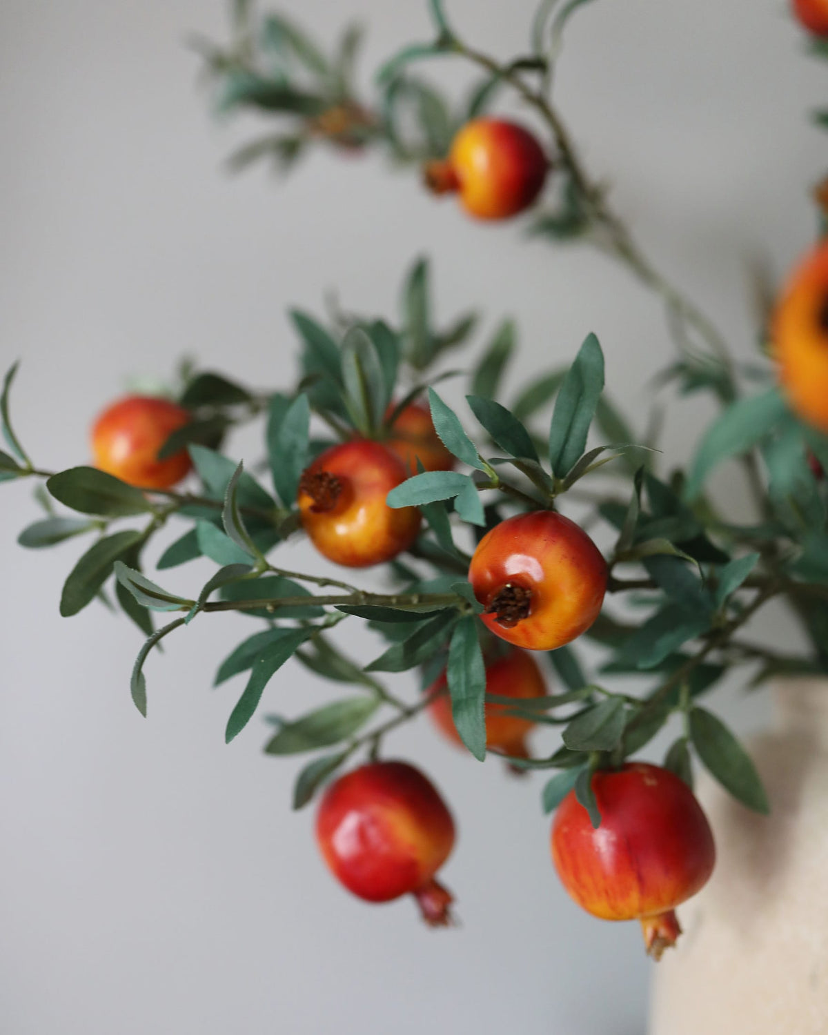 Artificial pomegranate branches with glossy red and orange fruits and green leaves, arranged against a soft light gray background.