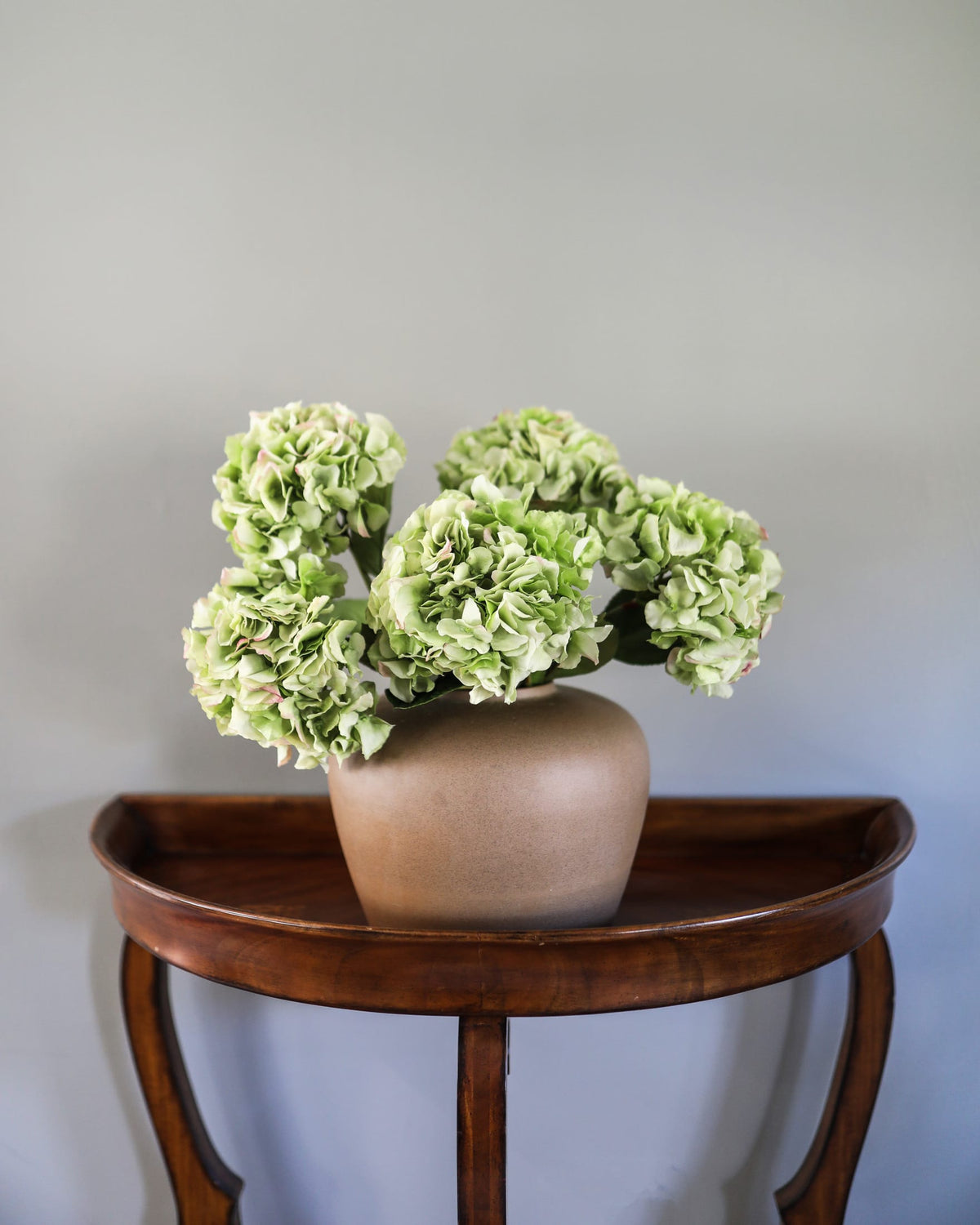 Artificial green real-touch hydrangeas arranged in a taupe vase on a dark wood console table.