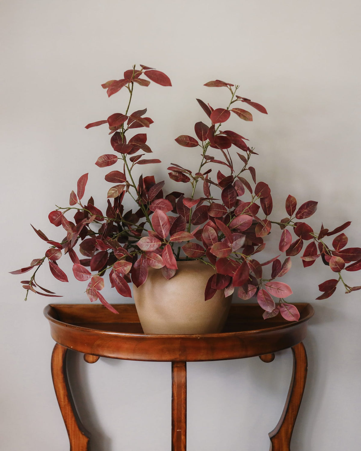An artificial potted plant with burgundy and pinkish-red variegated smoketree leaves, displayed in a beige ceramic pot on a curved wooden side table against a plain light gray wall.