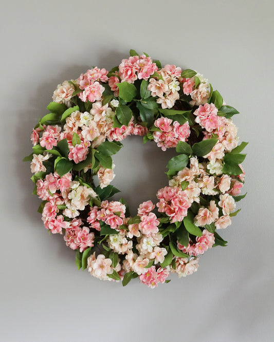 A circular floral wreath made of pink and cream blossoms with green leaves, photographed against a plain light gray wall.