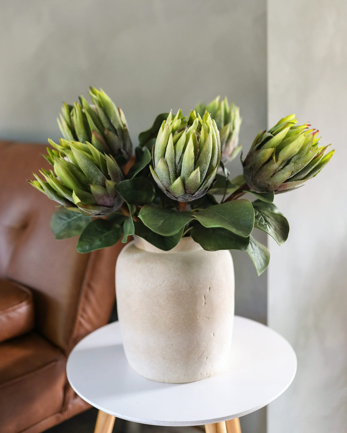 Faux green protea arrangement in a white vase on a small round table with a blurred background