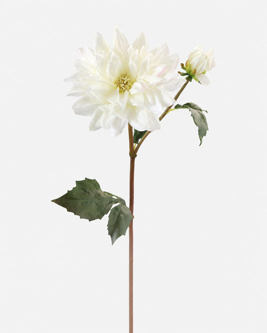 A single white faux dahlia flower stem with green leaves and a small bud, photographed against a plain light gray background.