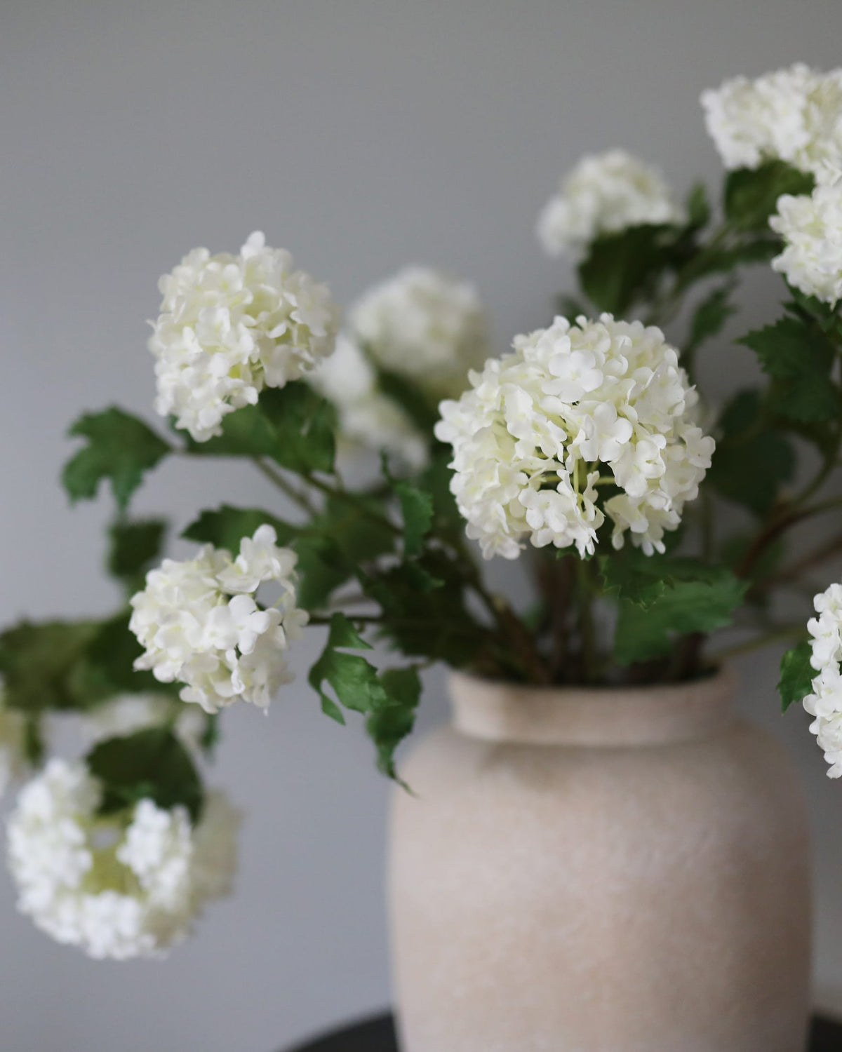 White faux snowball flower stems with green leaves arranged in a textured beige ceramic vase against a soft gray background.