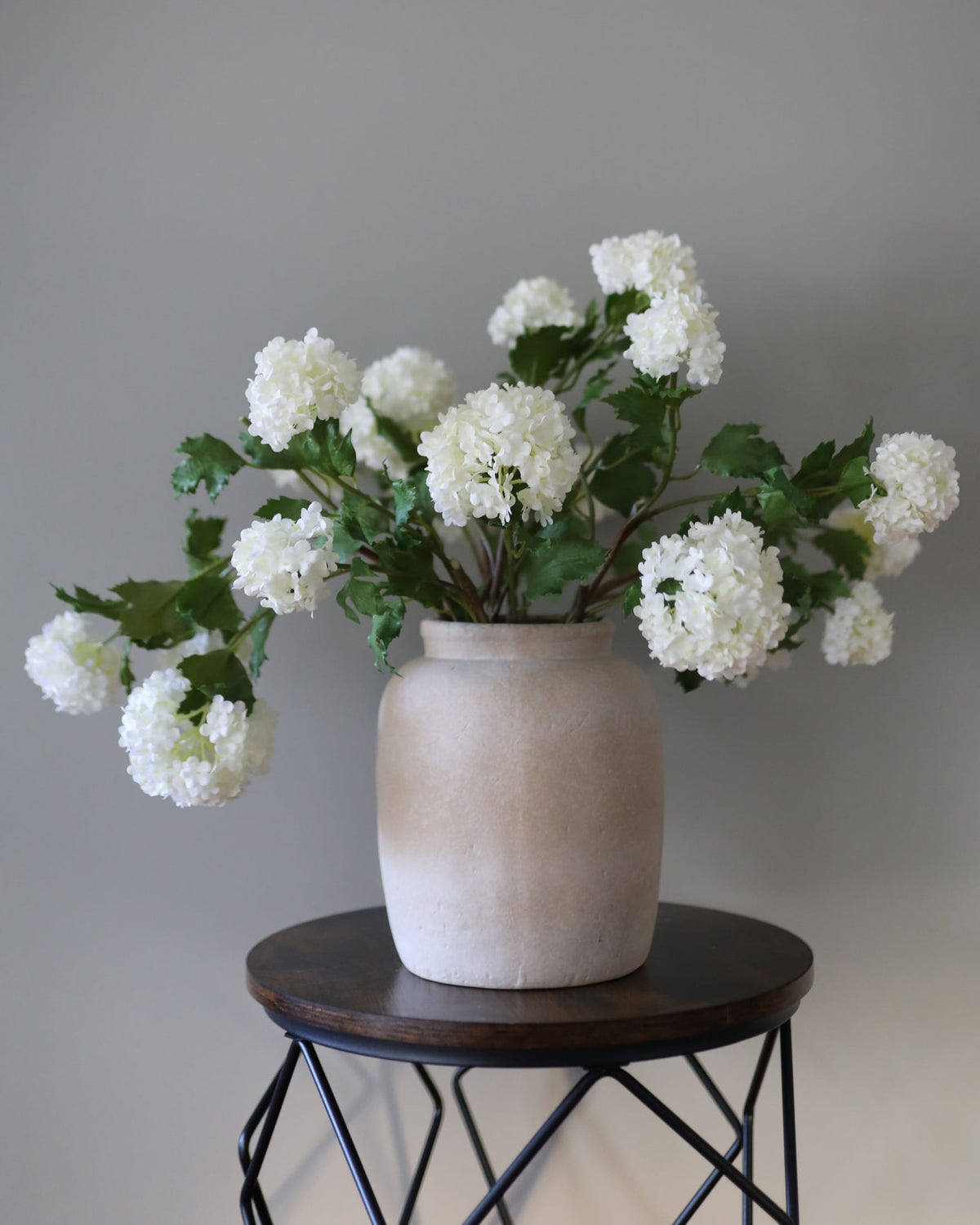 White faux snowball artificial flower arrangement in a textured beige ceramic vase, displayed on a dark wood and metal side table against a neutral gray wall.